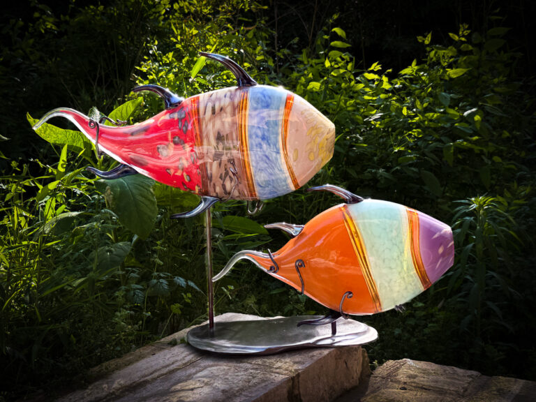 Colorful glass fish sculptures on metal stands displayed outdoors among green foliage.