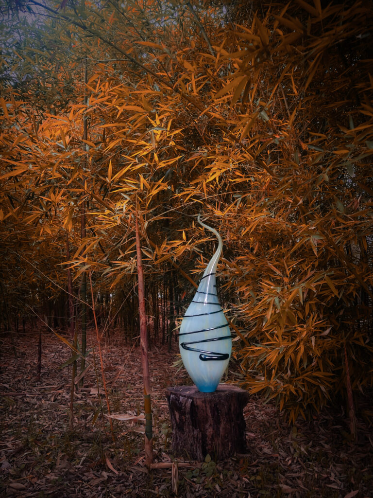 Blue spiral glass sculpture perched on a weathered tree stump among dense orange-banana bamboo leaves at dusk, creating a warm, glowing garden scene.
